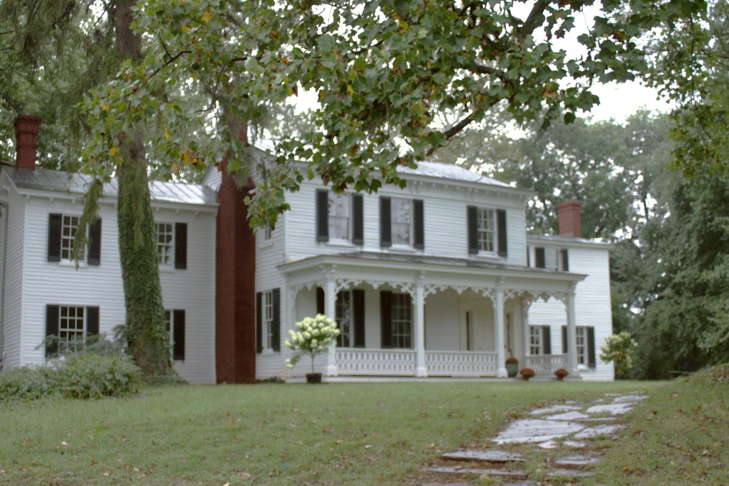 A white clapboard farmhouse with a decorated porch and brick chimneys under summer trees in Warrenton